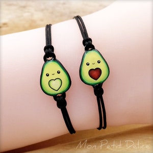 Puede incluir: Dos pulseras de amistad negras con abalorios de aguacate. Cada aguacate tiene una cara sonriente y un corazón en el centro. Un corazón es rojo y el otro es verde.