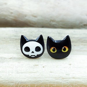 May include: A pair of black cat-shaped stud earrings. One earring features a white skull with a black mouth and eyes. The other earring has black eyes with orange pupils.