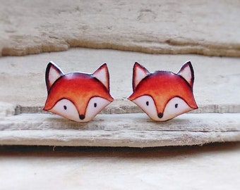 Puede incluir: Un par de pendientes con forma de zorro con pelaje naranja, vientre blanco y ojos negros. Los pendientes están sobre una superficie de madera.