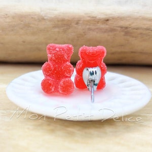 Könnte beinhalten: Ein Paar rote Gummibärchen-Ohrstecker auf einem weißen Teller. Die Ohrringe sind aus Harz und haben eine glänzende Oberfläche.