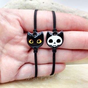 May include: Two black friendship bracelets with charms. One charm is a black cat with yellow eyes. The other charm is a white skull with black eyes.
