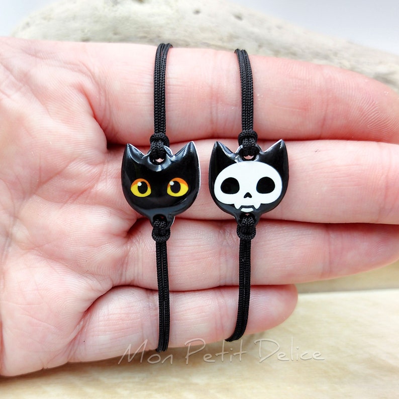 May include: Two black friendship bracelets with charms. One charm is a black cat with yellow eyes. The other charm is a white skull with black eyes.