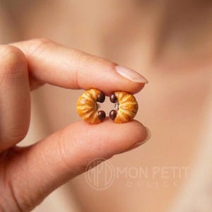 Tiny golden croissant stud earrings held between fingers to show small size reference.