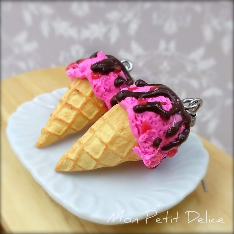 May include: A pair of pink and chocolate ice cream cone earrings. The earrings are made of polymer clay and feature a waffle cone with pink ice cream and chocolate sauce.