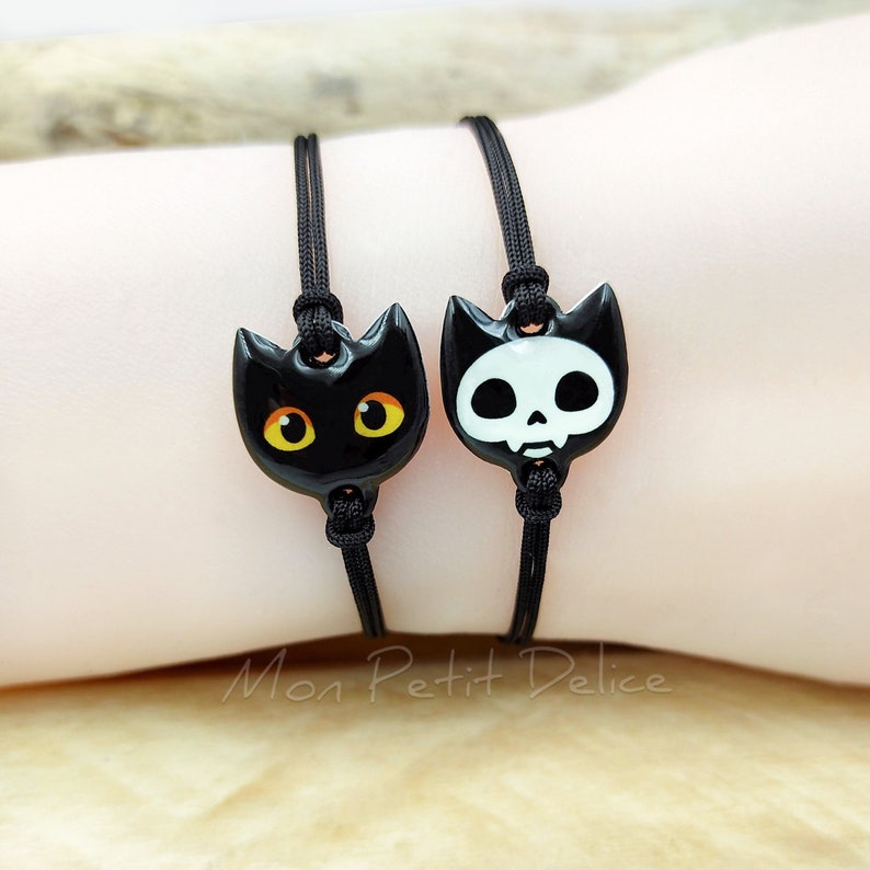 May include: Two black cord bracelets with charms. One charm is a black cat with yellow eyes. The other charm is a white skull with black details.