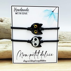 May include: Two black elastic bracelets with a black cat charm and a black and white skull charm. The bracelets are displayed on a white card with the text "Handmade with Love" and "Mon petit delice" with the website etsy.com/shop/monpetitdelice.