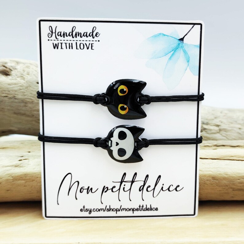 May include: Two black elastic bracelets with a black cat charm and a black and white skull charm. The bracelets are displayed on a white card with the text "Handmade with Love" and "Mon petit delice" with the website etsy.com/shop/monpetitdelice.