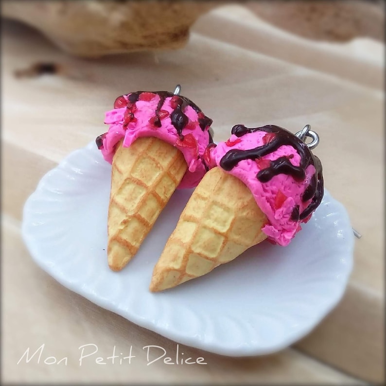 May include: A pair of pink and white ice cream cone earrings with chocolate sauce and sprinkles. The earrings are on a white plate with the text "Mon Petit Delice" on the bottom.