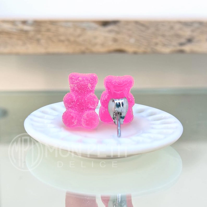 May include: A pair of bright pink gummy bear stud earrings. The earrings are shaped like gummy bears and have a silver post. The earrings are displayed on a white, round plate.