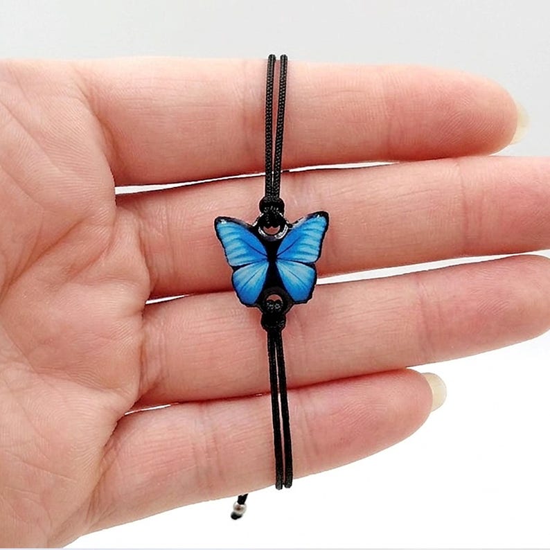 Blue monarch butterfly  bracelet with thick waterproof resin coating. Handcrafted jewelry featuring a sliding knot adjustable cord and anti-tarnish steel balls. The image shows the size in the model's hand.
