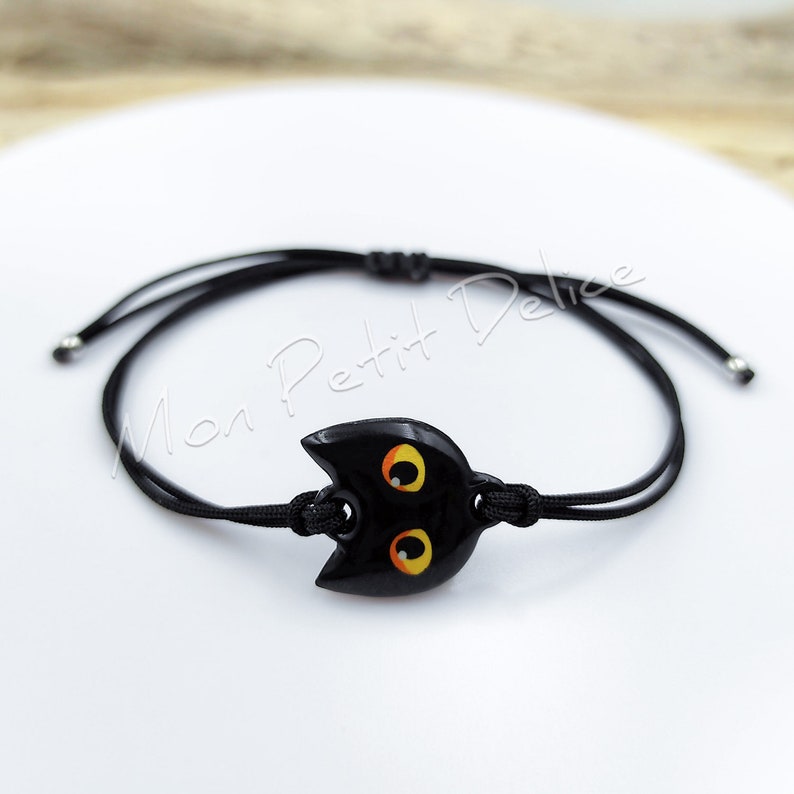 May include: Black adjustable bracelet with a black cat charm featuring yellow eyes. The charm is made of polymer clay.