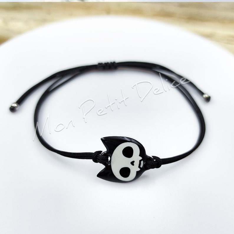 May include: Black adjustable bracelet with a black and white skull charm. The charm has a cat-like shape with ears and a white skull face.