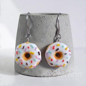 White donut earrings with colored sprinkles on a gray cement base. Miniature food jewelry.