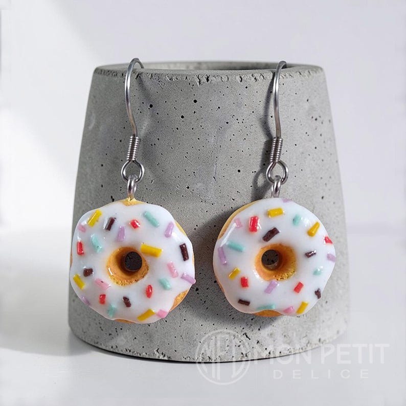 White donut earrings with colored sprinkles on a gray cement base. Miniature food jewelry.