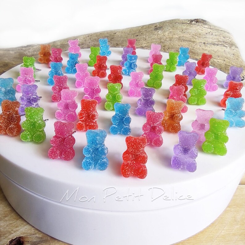 May include: A selection of multi-coloured gummy bear sweets, including red, blue, green, purple, and pink. The bears are arranged on a white surface, with the text "Mon Petit Delice" visible. The sweets are coated in sugar.