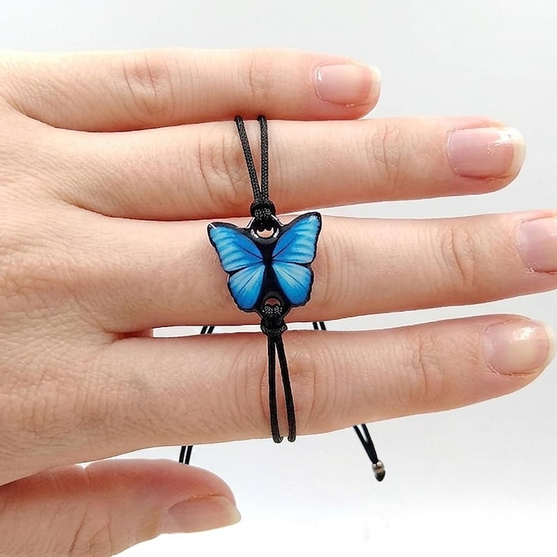 Blue and black butterfly charm bracelet with thick waterproof resin coating. Handcrafted jewelry featuring a sliding knot adjustable cord and anti-tarnish steel balls. Ideal gift for butterfly lovers. The image shows the size in the model's hand.