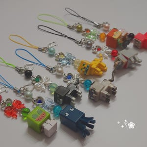 May include: A collection of seven phone charms featuring blocky, pixelated animal and creature figures in various colors including green, blue, black, tan, yellow, and orange. Each charm includes a colorful cord, metallic heart charms, and beads.