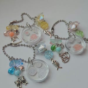 May include: Three clear resin charms with different colored beads and charms attached. Each charm features a small, white, cartoon-like image of a fish inside. The charms are attached to silver chains.