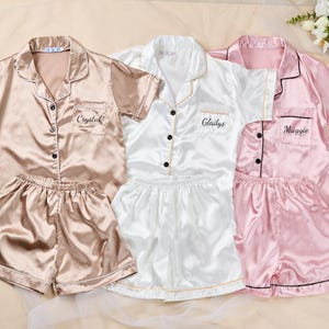 K&ouml;nnte beinhalten: Drei Sets Satin-Pyjamas in Champagner, Wei&szlig; und Rosa. Jedes Set enth&auml;lt ein kurz&auml;rmeliges Button-up-Shirt und passende Shorts. Die Hemden sind mit den Namen "Crystal", "Gladys" und "Maggie" personalisiert.