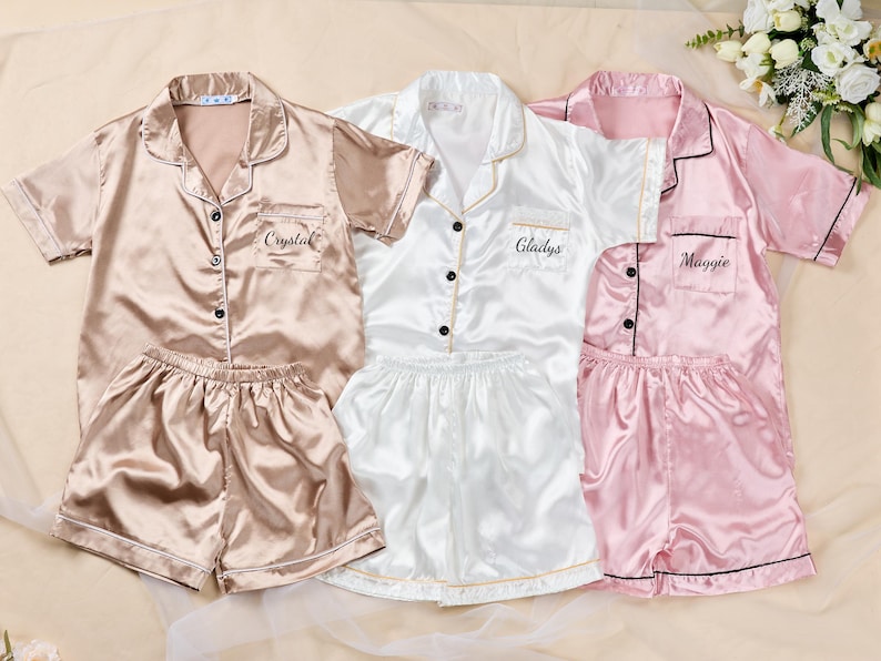 K&ouml;nnte beinhalten: Drei Sets Satin-Pyjamas in Champagner, Wei&szlig; und Rosa. Jedes Set enth&auml;lt ein kurz&auml;rmeliges Button-up-Shirt und passende Shorts. Die Hemden sind mit den Namen "Crystal", "Gladys" und "Maggie" personalisiert.