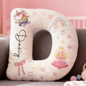Op de afbeelding: Een crèmekleurig kussen in de vorm van de letter "D" met geborduurde details. Het heeft een eenhoorn, regenboog, prinses, sterren, harten en een strik. De naam "Daisy" is in script geschreven. Een babyspeelgoed staat op de voorgrond.