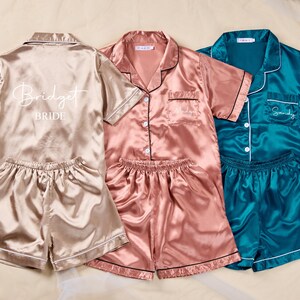K&ouml;nnte beinhalten: Drei Sets Satin-Pyjamas in Champagner, Rosa und T&uuml;rkis. Das Champagner-Set hat "Bridget Bride" auf dem R&uuml;cken aufgedruckt. Das rosafarbene Set hat einen Namen auf der Tasche gestickt. Das t&uuml;rkisfarbene Set hat "Sandy" auf der Tasche gestickt. Jedes Set enth&auml;lt ein kurz&auml;rmeliges Oberteil und Shorts.