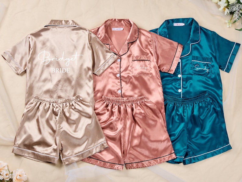 K&ouml;nnte beinhalten: Drei Sets Satin-Pyjamas in Champagner, Rosa und T&uuml;rkis. Das Champagner-Set hat "Bridget Bride" auf dem R&uuml;cken aufgedruckt. Das rosafarbene Set hat einen Namen auf der Tasche gestickt. Das t&uuml;rkisfarbene Set hat "Sandy" auf der Tasche gestickt. Jedes Set enth&auml;lt ein kurz&auml;rmeliges Oberteil und Shorts.