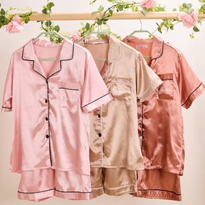 K&ouml;nnte beinhalten: Drei Sets Satin-Pyjamas in Rosa, Champagner und Koralle, h&auml;ngen an einem Holzgestell. Jedes Set enth&auml;lt ein kurz&auml;rmeliges Button-Down-Hemd und Shorts, die mit schwarzen Paspeln versehen sind. Die Hemden sind mit Namen personalisiert.