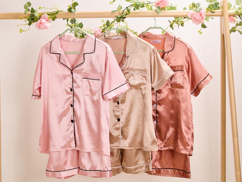 K&ouml;nnte beinhalten: Drei Sets Satin-Pyjamas in Rosa, Champagner und Koralle, h&auml;ngen an einem Holzgestell. Jedes Set enth&auml;lt ein kurz&auml;rmeliges Button-Down-Hemd und Shorts, die mit schwarzen Paspeln versehen sind. Die Hemden sind mit Namen personalisiert.
