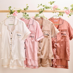 K&ouml;nnte beinhalten: Vier Sets Satin-Pyjamas in Wei&szlig;, Rosa, Beige und Koralle, jeweils mit personalisiertem Namen auf der Tasche gestickt. Die kurz&auml;rmeligen Hemden und Shorts h&auml;ngen an einer mit Blumen geschm&uuml;ckten Holzstange.
