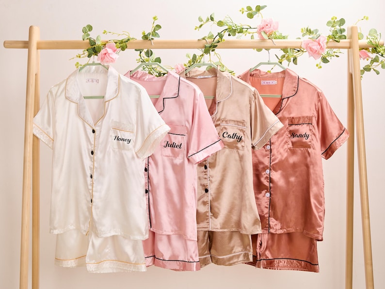 K&ouml;nnte beinhalten: Vier Sets Satin-Pyjamas in Wei&szlig;, Rosa, Beige und Koralle, jeweils mit personalisiertem Namen auf der Tasche gestickt. Die kurz&auml;rmeligen Hemden und Shorts h&auml;ngen an einer mit Blumen geschm&uuml;ckten Holzstange.
