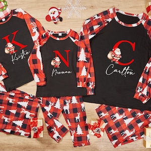 Pode incluir: Pijamas de Natal combinando em xadrez búfalo vermelho e preto. As blusas de manga comprida têm uma grande inicial e nome, com um gráfico do Papai Noel. As calças apresentam um padrão correspondente com renas e árvores de Natal.