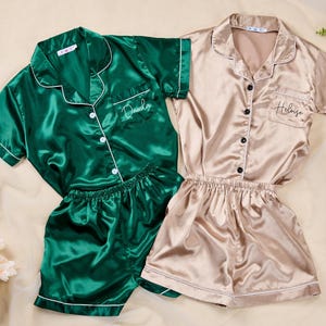 K&ouml;nnte beinhalten: Zwei Sets Satin-Pyjamas. Ein Set ist smaragdgr&uuml;n mit wei&szlig;er Paspelierung und dem Namen "Danila" auf der Tasche gestickt. Das andere Set ist champagnerfarben mit schwarzen Kn&ouml;pfen und dem Namen "Heloise" auf der Tasche gestickt. Beide Sets beinhalten ein kurz&auml;rmeliges Hemd mit Kn&ouml;pfen und Shorts.