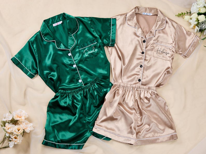 K&ouml;nnte beinhalten: Zwei Sets Satin-Pyjamas. Ein Set ist smaragdgr&uuml;n mit wei&szlig;er Paspelierung und dem Namen "Danila" auf der Tasche gestickt. Das andere Set ist champagnerfarben mit schwarzen Kn&ouml;pfen und dem Namen "Heloise" auf der Tasche gestickt. Beide Sets beinhalten ein kurz&auml;rmeliges Hemd mit Kn&ouml;pfen und Shorts.