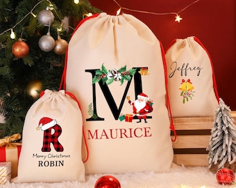 Puede incluir: Tres bolsas de regalo navideñas beige con cordones rojos. Una bolsa presenta una gran letra negra "M" con acebo y un gráfico de Papá Noel, el nombre "Maurice" debajo. Otra bolsa tiene una "R" a cuadros con un gorro de Papá Noel y el texto "Merry Christmas ROBIN". La tercera bolsa tiene el nombre de "Jeffrey".