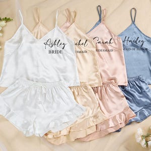 Könnte beinhalten: Satin-Pyjama-Sets in Weiß, Beige, Pfirsich und Blau. Jedes Set enthält ein Camisole-Top und Shorts mit Rüschen. Die Oberteile sind mit Namen und Titeln wie "Braut" und "Brautjungfer" personalisiert. Ein Strauß weißer Blumen ist im Hintergrund.