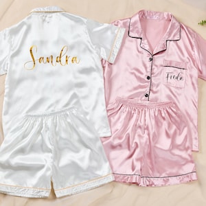 K&ouml;nnte beinhalten: Zwei Sets aus Satin-Pyjamas. Ein Set ist wei&szlig; mit goldenem Text, der "Sandra" auf dem R&uuml;cken zeigt. Das andere Set ist rosa mit schwarzem Besatz und dem Namen "Freda" auf der Tasche. Beide Sets beinhalten ein Oberteil mit Kn&ouml;pfen und Shorts.