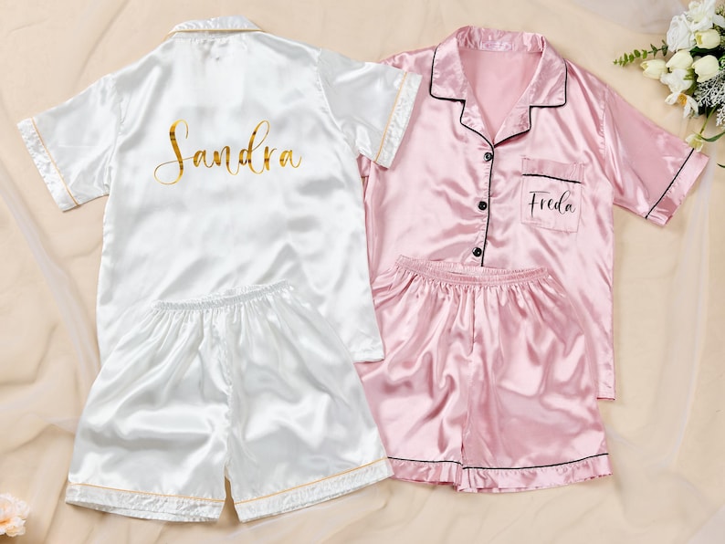 K&ouml;nnte beinhalten: Zwei Sets aus Satin-Pyjamas. Ein Set ist wei&szlig; mit goldenem Text, der "Sandra" auf dem R&uuml;cken zeigt. Das andere Set ist rosa mit schwarzem Besatz und dem Namen "Freda" auf der Tasche. Beide Sets beinhalten ein Oberteil mit Kn&ouml;pfen und Shorts.