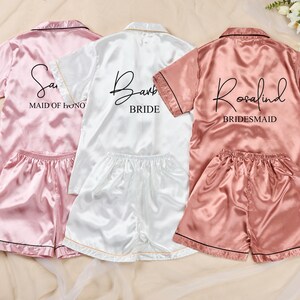 K&ouml;nnte beinhalten: Drei Satin-Pyjama-Sets in Rosa, Wei&szlig; und Ros&eacute;. Jedes Set enth&auml;lt ein kurz&auml;rmeliges Oberteil und Shorts. Das rosa Set tr&auml;gt die Aufschrift "Sam" und "Maid of Honor". Das wei&szlig;e Set tr&auml;gt die Aufschrift "Barb" und "Bride". Das rosafarbene Set tr&auml;gt die Aufschrift "Rosalind" und "Bridesmaid".