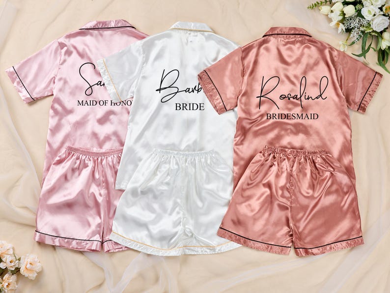 K&ouml;nnte beinhalten: Drei Satin-Pyjama-Sets in Rosa, Wei&szlig; und Ros&eacute;. Jedes Set enth&auml;lt ein kurz&auml;rmeliges Oberteil und Shorts. Das rosa Set tr&auml;gt die Aufschrift "Sam" und "Maid of Honor". Das wei&szlig;e Set tr&auml;gt die Aufschrift "Barb" und "Bride". Das rosafarbene Set tr&auml;gt die Aufschrift "Rosalind" und "Bridesmaid".