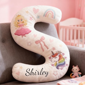 May include: A cream-colored, letter "S" shaped pillow with princess, unicorn, and rainbow designs. The pillow has the name "Shirley" printed on it. The pillow is decorated with hearts and stars.