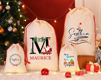 Pode incluir: Uma coleção de sacos de presente de Natal creme com cordões vermelhos. Os sacos apresentam designs festivos, incluindo uma coroa com o nome "Sophia", uma árvore de Natal com o nome "Sophia" e uma ilustração do Papai Noel com o nome "Sampson".