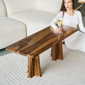 Könnte beinhalten: Ein rechteckiger Couchtisch aus Holz mit einer satten, warmen Holzmaserung. Der Tisch hat ein einzigartiges Design mit einer hellen Intarsie. Der Tisch wird von vier stabilen, ausgestellten Beinen getragen. Der Tisch ist ca. 122 cm lang.
