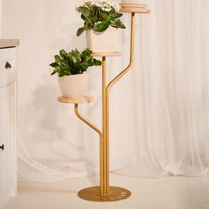 Może przedstawiać: Złoty metalowy stojak na rośliny z trzema drewnianymi półkami, z których każda mieści białą ceramiczną donicę z inną kwitnącą rośliną. Stojak ma okrągłą podstawę i konstrukcję przypominającą drzewo.