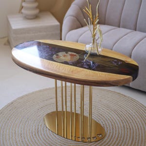 May include: Oval coffee table with a wooden top inlaid with a floral design and a dark resin border. The table has gold-colored metal legs and a round base. A small glass vase with dried plants sits on the table.