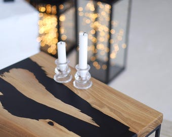 Op de afbeelding: Een houten tafel met een zwart epoxyharsontwerp. Twee doorzichtige glazen kandelaars met witte kaarsen staan op de tafel. Twee zwarte lantaarns met lichtslingers staan op de achtergrond.
