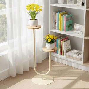 May include: A cream-coloured metal plant stand with two tiers and light wood shelves. Each shelf holds a white pot with yellow flowers. The stand has a circular base and a curved design, positioned near a window with white curtains.