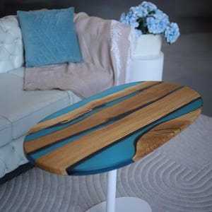 May include: Oval coffee table with a wooden top and a white base. The tabletop has a wood grain design with blue resin inlays. The table is set on a light grey rug. The table's dimensions are approximately 100 cm in length.