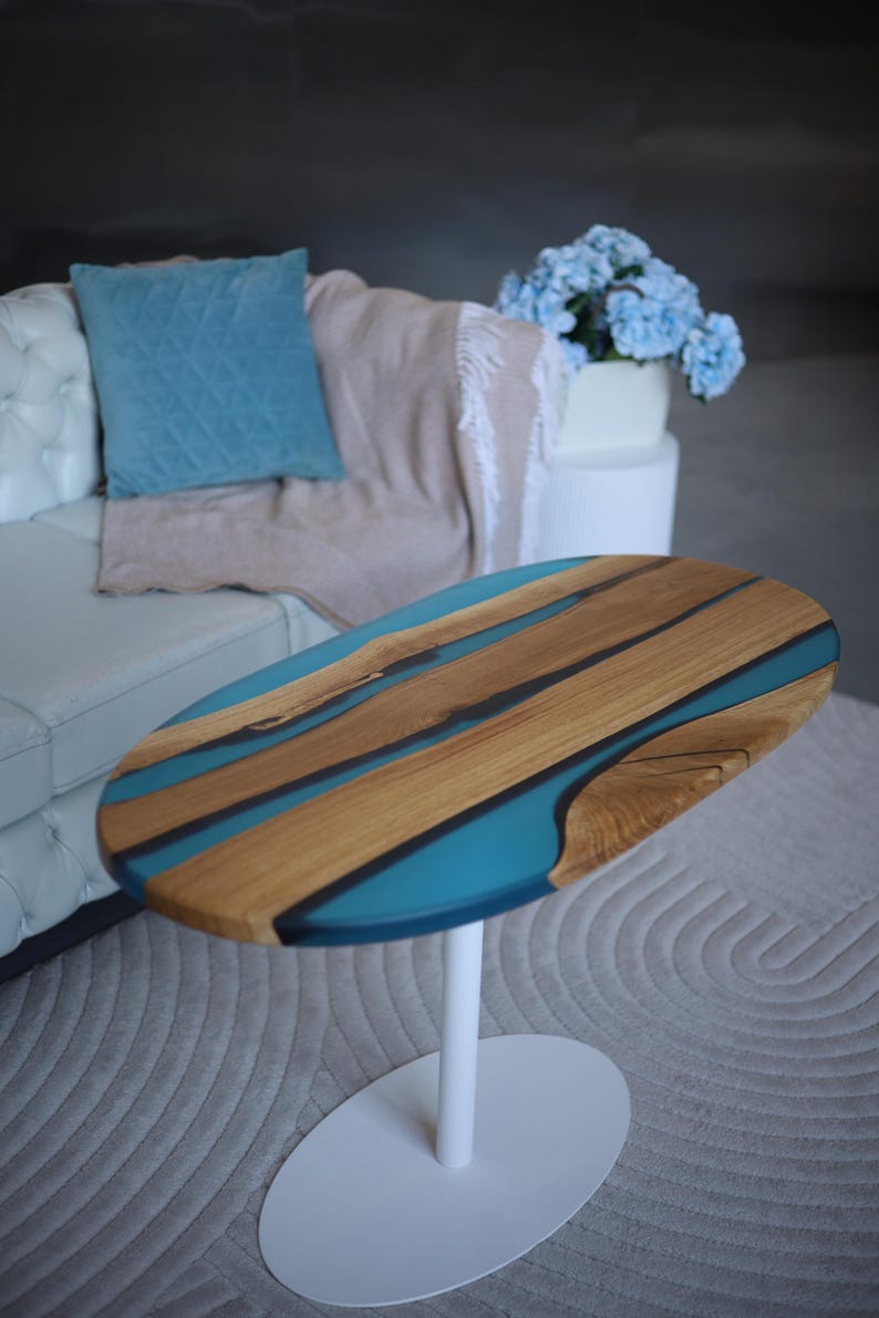 May include: Oval coffee table with a wooden top and a white base. The tabletop has a wood grain design with blue resin inlays. The table is set on a light grey rug. The table's dimensions are approximately 100 cm in length.