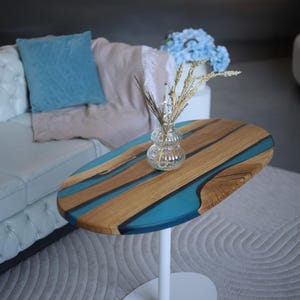 May include: An oval side table with a wooden top and blue resin river design. The table is supported by a white pedestal base. A glass vase with dried flowers sits on the table. The table is placed on a grey rug.